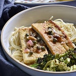 Tofu Piccata on a plate of spaghetti and roasted broccoli, with a platter of tofu piccata in the back