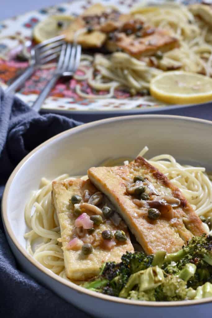 Easy Tofu Piccata [with lemon and capers]  on a plate of spaghetti and roasted broccoli, with a platter of tofu piccata in the back